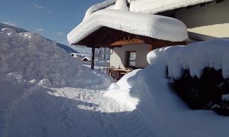 A snowy path leads to a house with a snow-covered roof. The sky is clear and blue, creating a winter atmosphere.