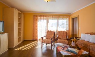 A bright living room with orange walls and large windows. It includes comfortable armchairs, a sofa, and a television.