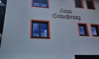 A building with the inscription "Haus Gamsprung". The facade is white with orange window frames and a well-kept garden in the foreground.