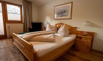 A cozy bedroom with a wooden bed and comfortable pillows. The room has large windows and a warm, inviting floor.