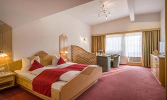 A bright and inviting room with a large bed and red accents. The room features a cozy seating area and large windows with a view outside.