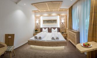 A cozy hotel room with a double bed and light wood details. Fresh fruit is placed on a table, and large windows with curtains create a pleasant atmosphere.