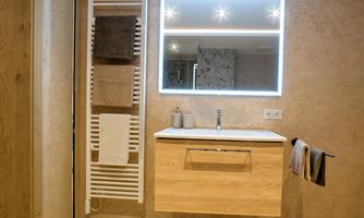A modern bathroom with a wooden sink and a lit mirror. Towels hang on a towel warmer and on a bar.