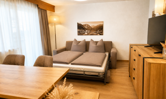 A cozy living room with a sofa bed and a wooden table.  
Large windows provide plenty of daylight and a warm atmosphere.