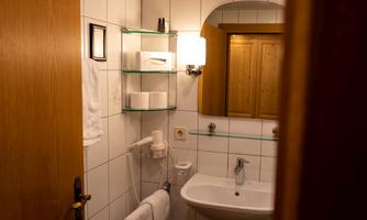 A small bathroom with white tiles and a sink. Towels and toiletries are arranged on shelves on the wall.