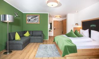A modern room with green painted walls and a double bed. There is a cozy couch and a few decorative cushions in a fresh design.