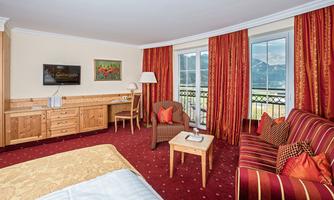 A cozy hotel room with wooden furniture and red-gold curtains. The room offers a view of the mountains through large windows.