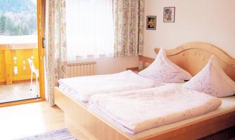 A bright bedroom with a double bed and large windows. In the background, there is a balcony overlooking the mountains.