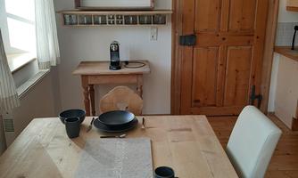 A cozy dining area with a wooden table and black plates. The atmosphere is bright and inviting with wooden details and windows.