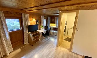 A cozy living space with wooden walls and a television. In the background, there is a small dining area and a bathroom.