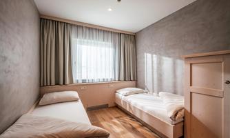 A simple bedroom with two single beds and a window. The walls are in a neutral color and the floor is made of wood.