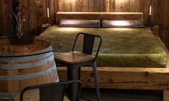 A cozy bedroom with a wooden bed and a green mattress. Next to the bed, there are two chairs and a table made from a wine barrel.