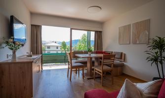 A modern living room with large windows and access to a balcony. The room is bright and welcoming, featuring a dining table and cozy furniture.