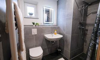 A modern bathroom with gray tiles and a shower. The decor is simple and functional, featuring a small sink and plenty of natural light.