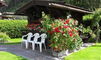 A cozy garden house surrounded by green bushes and colorful flowers. In front of the house, three white chairs are situated on a paved pathway.