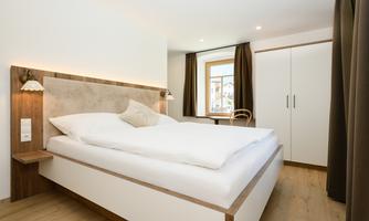 A modern bedroom with a comfortable bed and a large window. The furnishings are simple and elegant, with warm wood and white tones.