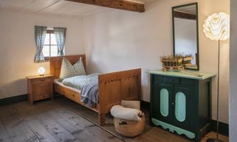A cozy bedroom with a single bed, a small dresser, and a decorative cabinet. The windows are adorned with checked curtains and the floor is made of wood.