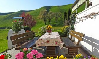 An inviting garden with a wooden table and benches, surrounded by colorful flowers. In the background, gentle green hills can be seen and the sky is clear.