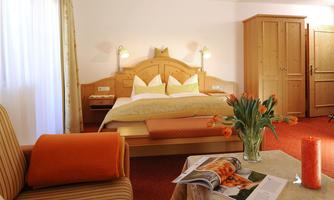 A cozy bedroom with a large bed, bright cushions, and a comfortable sofa. Fresh flowers and a candle are on the table.