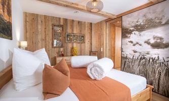 A cozy bedroom with a wooden bed and decorative pillows. The walls are made of wood, and a large photo wall displays a landscape.