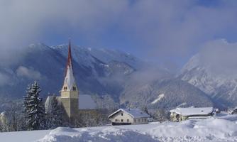 A picturesque winter landscape with snow-covered mountains. In the foreground stands a church with a red tower and some attractive houses.