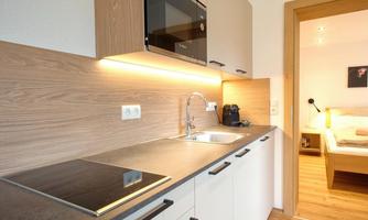 A modern kitchen with wooden fronts and an elegant workspace. In the background, a cozy bedroom is visible.