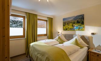 A cozy bedroom with two beds and green bedding. Large windows offer a view of nature, and a beautiful mural complements the atmosphere.
