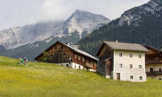 A picturesque mountain landscape with green meadows and traditional wooden houses. In the background, majestic mountains rise under a cloudy sky.