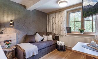 A cozy living room with a sofa, cushions, and a table. Large windows let in plenty of light and offer a view outside.