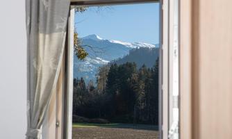 The view from the window shows snow-covered mountains and a clear blue sky. In the foreground, trees and a frost-resistant field are visible.