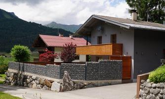 A modern house with wooden accents and a beautiful garden. In the background, gentle hills and green trees can be seen.