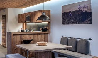 A modern kitchen with wooden shelves and a dining area. A picture of a mountain landscape hangs on the wall.