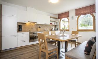 A modern kitchen with bright furniture and large windows. The dining table is equipped with wooden chairs and decorative elements.