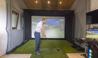 A man is playing golf in an indoor golf room with a virtual screen. The room is equipped with a net and artificial grass.