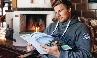 A man sits relaxed in a cozy room and reads a travel brochure. In the background, a fireplace is burning, creating a warm atmosphere.