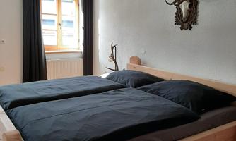 A cozy bedroom with a wooden bed and two black pillows. On the wall hang deer antlers and windows with curtains.