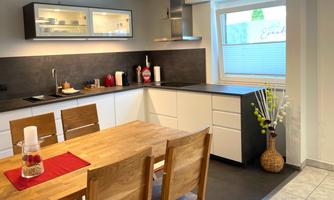 A modern kitchen with a wooden dining table and stylish chairs. The walls are light and there are large windows that let in plenty of light.