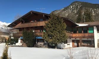 A cozy, old wooden house in the snow with a large balcony. In the background, mountains and a clear blue sky can be seen.