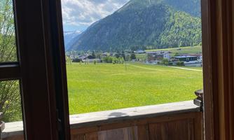 A view from a wooden window onto a green meadow and a mountain in the background. The sky is partly cloudy and radiates a tranquil atmosphere.