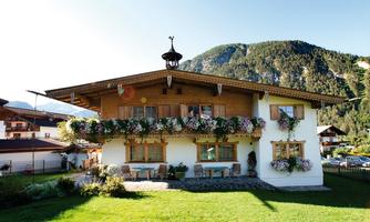 A cozy wooden house with a balcony and blooming plants. In the background, there are green mountains and a blue sky.