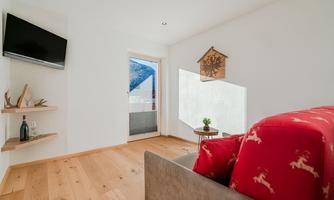 A modern living room with a TV on the wall and a cozy sofa. Large windows provide a view of the surroundings and there is a small plant on a table.