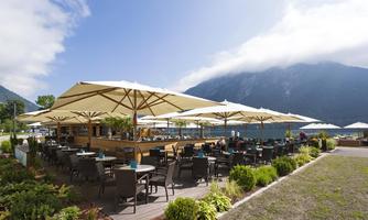 An inviting café by the lake with sun umbrellas and plenty of seating. In the background, majestic mountains and a clear blue sky can be seen.