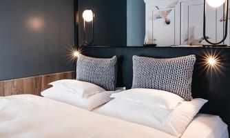 A modern bedroom with a double bed and stylish pillows. The walls are in dark colors, and the lighting creates a cozy atmosphere.