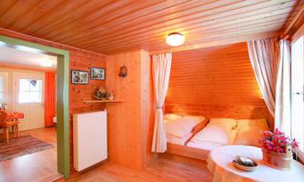 A cozy bedroom with wood paneling and two beds. The room is bright and welcoming with windows and a small table.