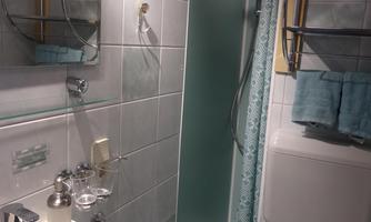 A small, brightly tiled bathroom with a shower and a sink. Towels in soft colors hang on a shelf.