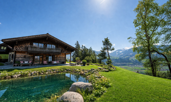 A magnificent wooden house surrounded by green grass and a peaceful landscape. In the foreground, there is a pond that reflects the surrounding nature.