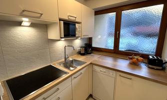 A modern kitchen with bright furniture and a window that lets in plenty of daylight. On the countertop, there are fruits and various kitchen appliances.