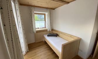 A simple room with a wooden bed and a window through which light falls. The walls are light and the floors are made of wood.