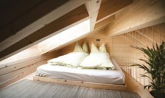A cozy room in the attic with a bed and green pillows. Bright wood walls and a plant create an inviting atmosphere.