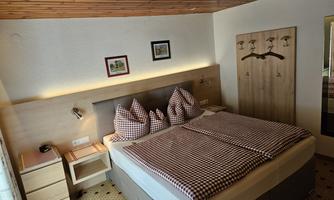 A cozy bedroom with a double bed and checkered pillows. The walls are adorned with pictures, and there is a clothes rack as well as a bedside table.